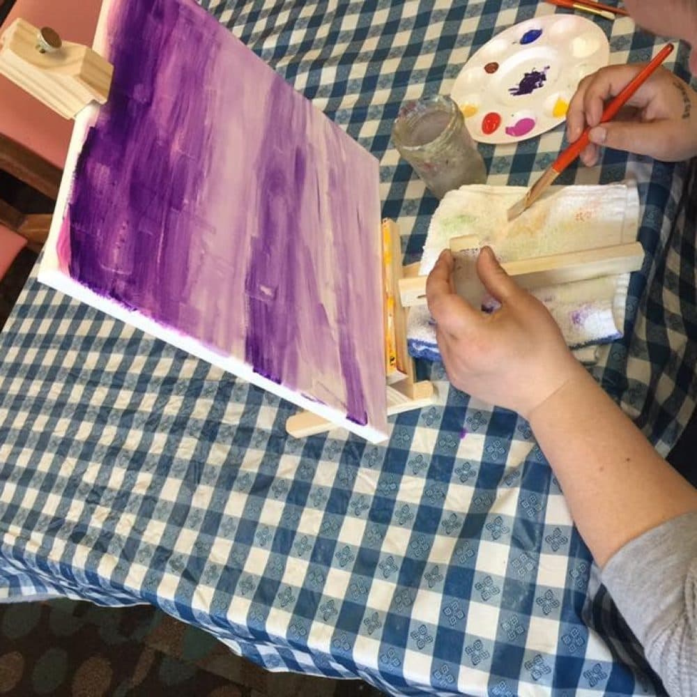 Painter painting a purple sunset on an easle that is sitting on a blue tablecloth.