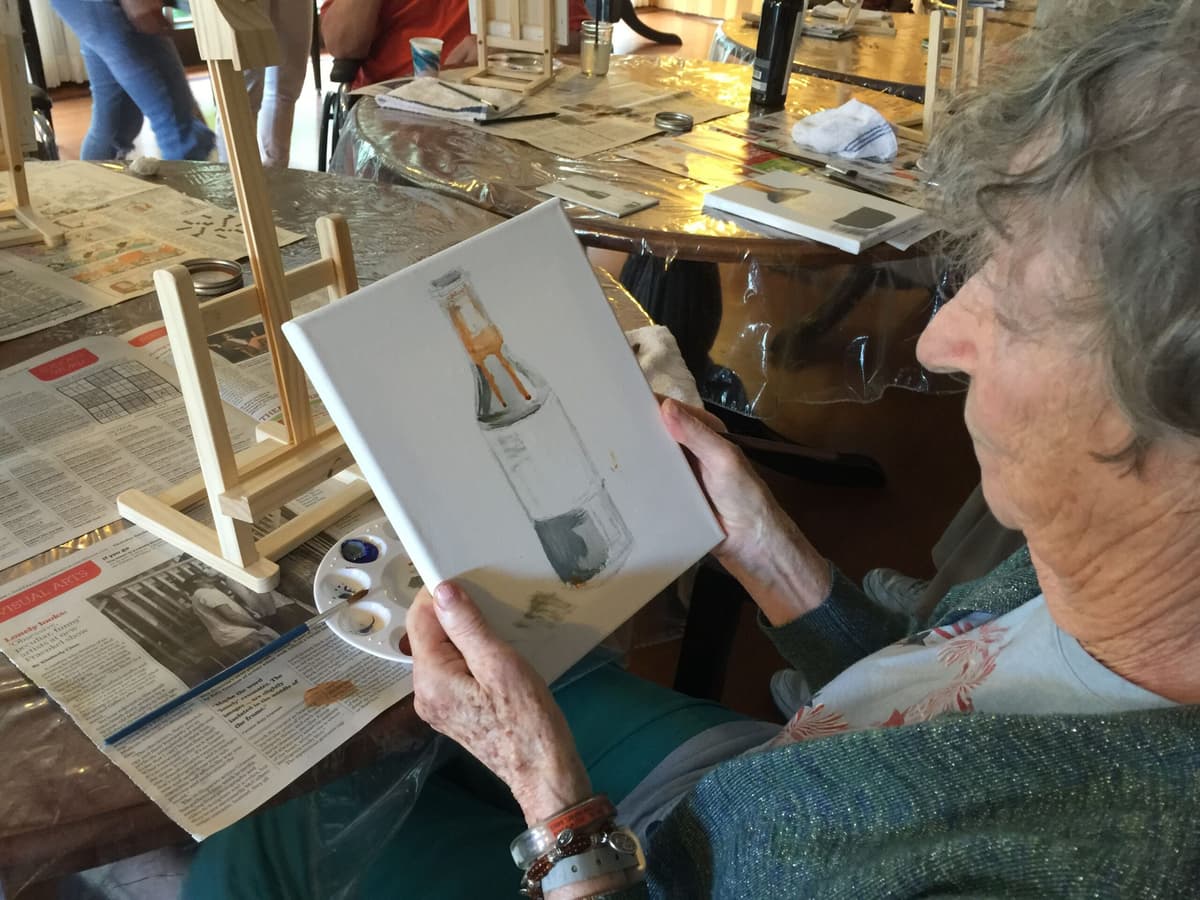 Elderly woman painting a wine bottle in water color paints canvas.